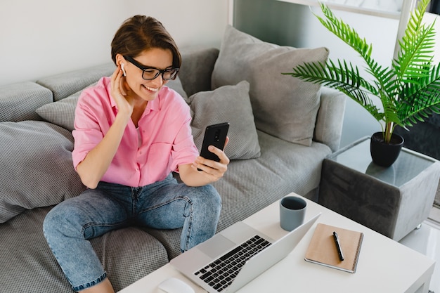 Aantrekkelijke lachende vrouw in roze shirt zittend ontspannen op de bank thuis aan tafel online werken op laptop vanuit huis