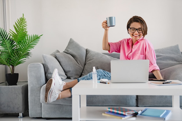 Aantrekkelijke lachende vrouw in roze shirt zittend ontspannen thee drinken op de bank thuis aan tafel online werken op laptop thuis