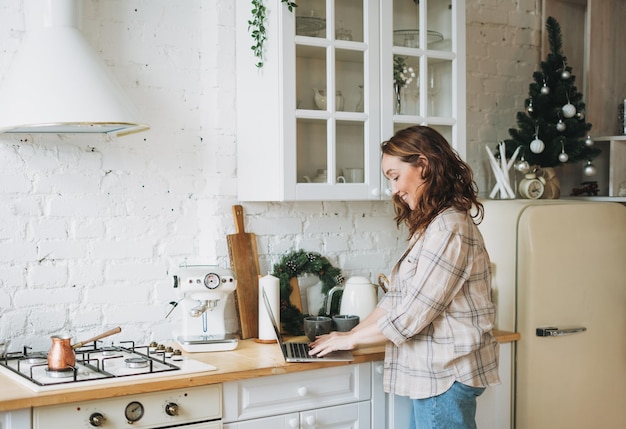 Aantrekkelijke lachende vrouw krullend haar in geruite hemd met behulp van laptop in lichte keuken