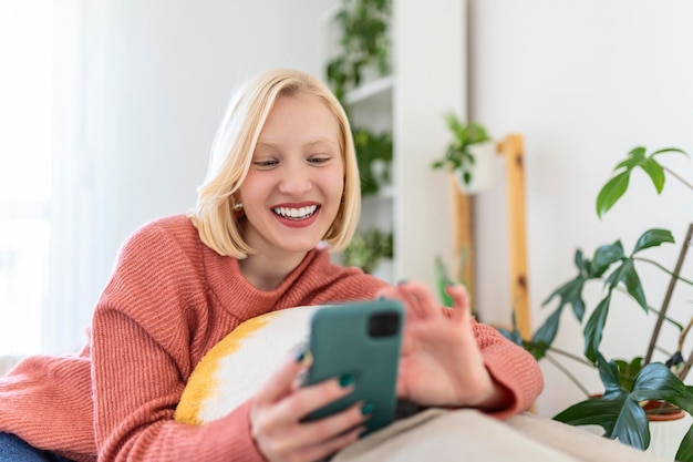 Aantrekkelijke lachende vrouw met behulp van slimme telefoon zittend op de bank thuis. Communicatie en gezelligheid concept.