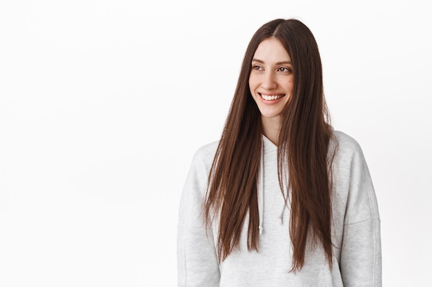 Aantrekkelijke lachende vrouw met lang bruin haar, hoofd opzij, kijkend naar logo met vrolijk gezicht, banner links lezend, staande in hoodie tegen witte muur