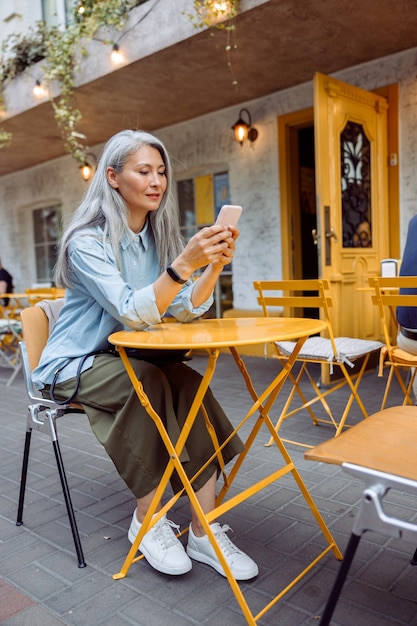 Aantrekkelijke langharige senior aziatische dame houdt mobiele telefoon vast aan een gele tafel
