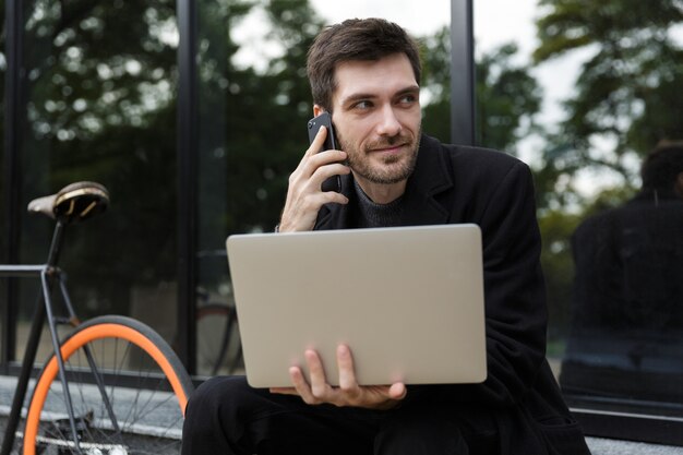 Aantrekkelijke man gekleed in jas zittend aan de straat, met behulp van laptopcomputer, praten op een mobiele telefoon