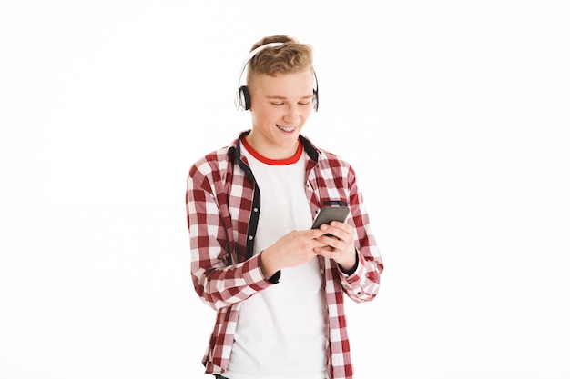 Aantrekkelijke man in casual t-shirt met beugels luisteren naar favoriete deuntje via draadloze hoofdtelefoons en met behulp van moderne gadget, geïsoleerd over witte muur