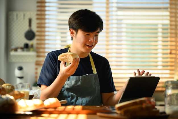 Aantrekkelijke man in schort met vers brood zittend aan eettafel in keuken en videogesprek via laptop