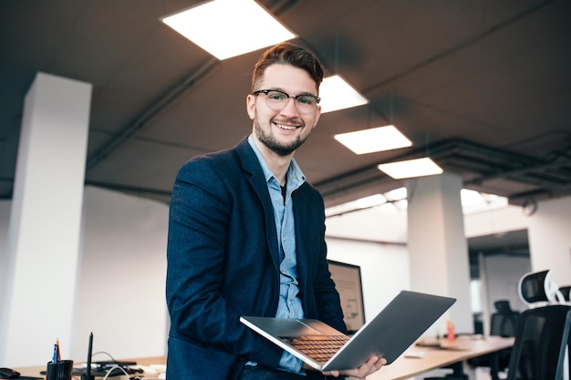 Aantrekkelijke man met bril staat in de buurt van de werkplek op kantoor. Hij draagt een blauw shirt, een donkere jas. Hij typt op laptop.