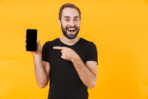 Aantrekkelijke man met een zwart basic t-shirt glimlachend en wijzend naar de mobiele telefoon geïsoleerd over een gele muur