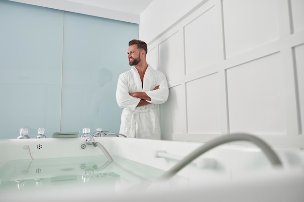 Aantrekkelijke man met gekruiste armen staat in de buurt van hydromassagebad in spasalon