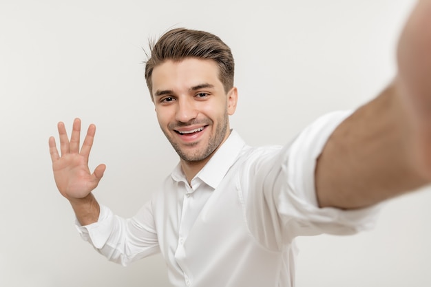 Foto aantrekkelijke man met wit overhemd die selfie of videogesprek maakt en hallo zegt, geïsoleerd over wit