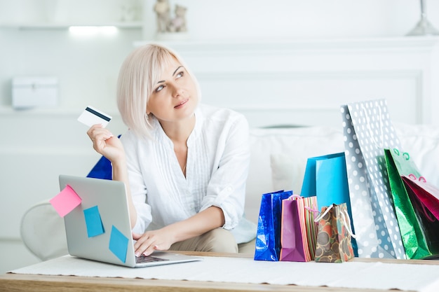 Aantrekkelijke medio volwassen vrouw die online shoppind doet. De vrouwelijke holdingscreditcard en zitting bij laptop met het winkelen doet binnen in zakken.