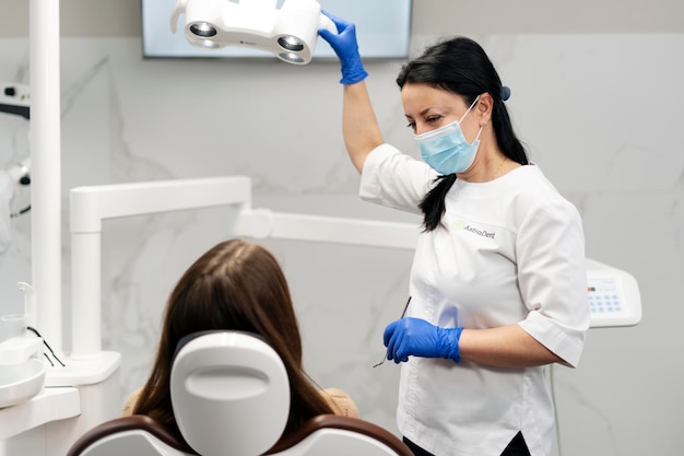 Aantrekkelijke mooie vrouwelijke tandarts die een beschermend masker en handschoenen draagt, praat met een vrouwelijke patiënt die in de tandartsstoel zit Het concept van tandheelkundige behandeling gezondheidszorg