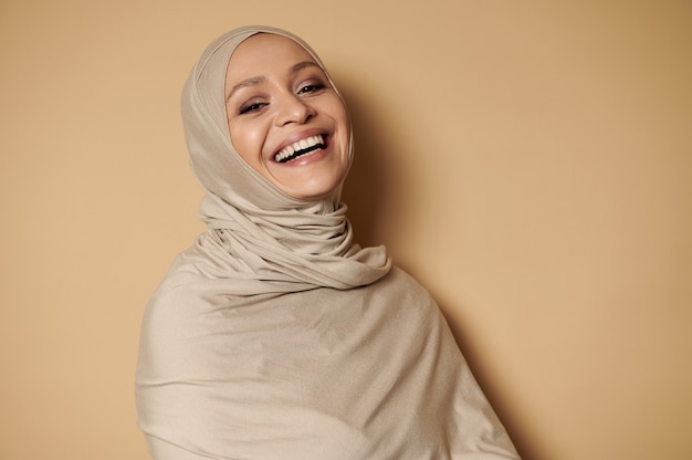 Aantrekkelijke moslimvrouw in hijab, lacht camera kijken tegen beige met kopie ruimte