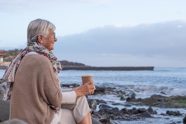 Aantrekkelijke ontspannen Kaukasische senior vrouw zittend op het kiezelstrand bij zonsondergang licht bewonderende horizon over water Glimlachende oudere dame met een koffiekopje in de hand