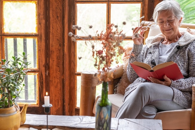 Aantrekkelijke ontspannen senior vrouw om thuis te zitten in een leunstoel die een boek leest Bejaarde lachende blanke vrouw geniet van leesplezier met een bril in de hand