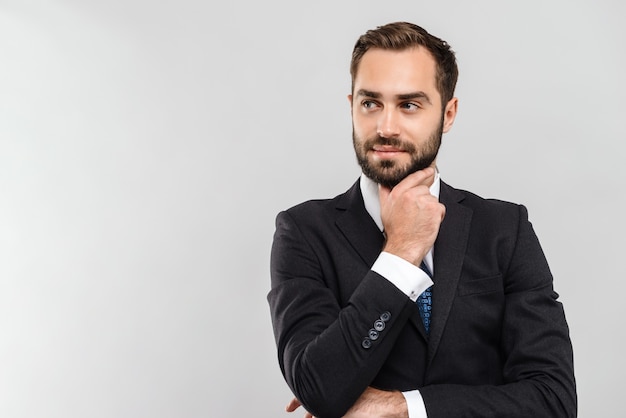 Aantrekkelijke peinzende jonge zakenman die een pak draagt dat geïsoleerd staat over een grijze muur