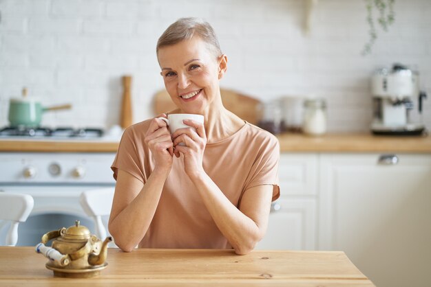 Aantrekkelijke rijpe vrouw begint de dag met koffie zittend aan houten tafel in de keuken