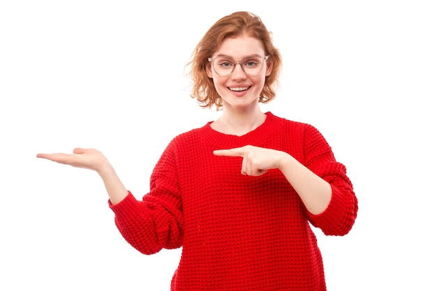 Aantrekkelijke roodharige meisje promotor in rode trui iets in de hand te houden demonstreren lege ruimte met opgewonden gezicht geïsoleerd op witte achtergrond