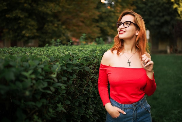 Aantrekkelijke roodharige vrouw met een bril draagt een rode blouse en jeansrok die zich voordeed in het groene park