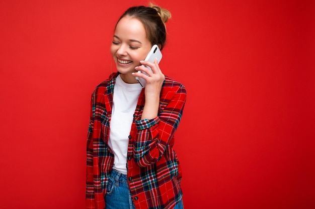 Aantrekkelijke schattige positieve jonge blonde vrouw staande geïsoleerd over rode muur dragen casual rood shirt en wit t-shirt praten op mobiele telefoon op zoek naar de kant.