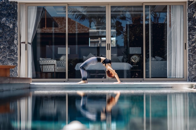 Aantrekkelijke schattige sportieve vrouw die sportkleding draagt en yoga doet naast het zwembad op de achtergrond van haar baan. Yogaconcept