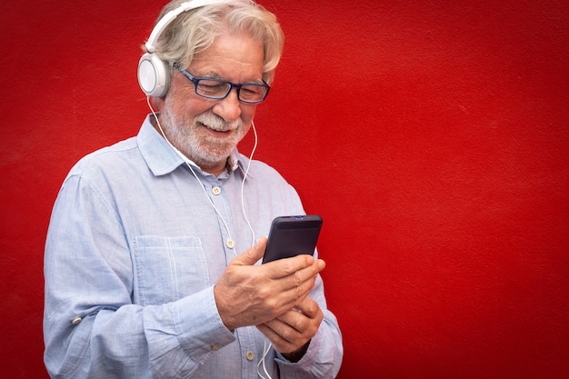 Aantrekkelijke senior bebaarde man staande op rode achtergrond met koptelefoon luisteren naar muziek van slimme telefoon. Kaukasische mensen glimlachen zorgeloos
