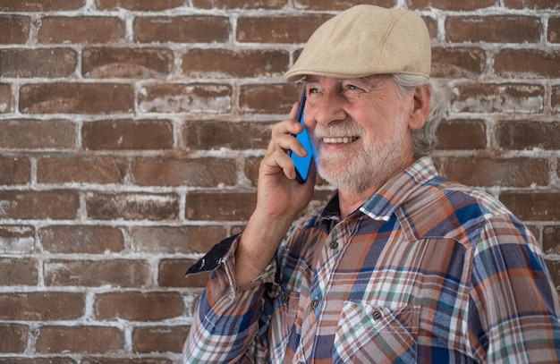 Aantrekkelijke senior man casual kleding staande tegen een bakstenen muur met behulp van telefoon en glimlachen.