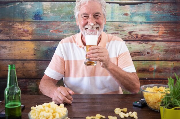 Aantrekkelijke senior man zit aan houten tafel met een glas blond bier, kijkend naar camera glimlachend