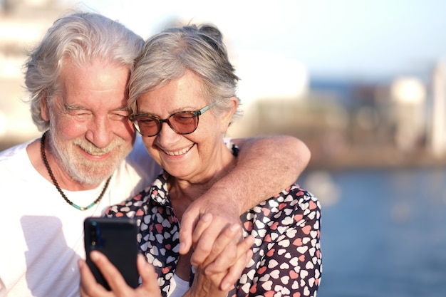 Aantrekkelijke senior paar glimlachend in de buitenlucht op zee met behulp van mobiele telefoon. kaukasisch paar witharig