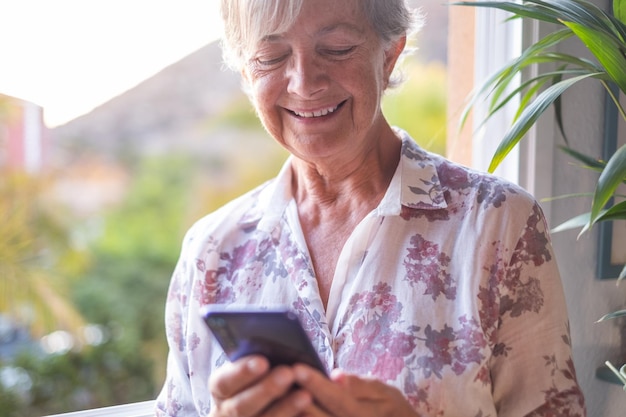 Aantrekkelijke senior vrouw bij het raam met mobiele telefoon tijdens het schrijven van een bericht. Glimlachende grijsharige senior genieten van technologie en sociaal