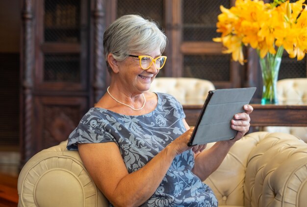 Aantrekkelijke senior vrouw zittend op de bank met behulp van digitale tablet. Glimlachende moderne gepensioneerde die geniet van een ontspannend moment