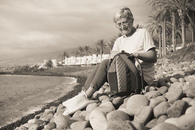 Aantrekkelijke senior vrouw zittend op een kiezelstrand op zee kijken in haar rugzak volwassen vrouw glimlacht terwijl het rusten genieten van vakantie Concept van vrijheid en gelukkig pensioen