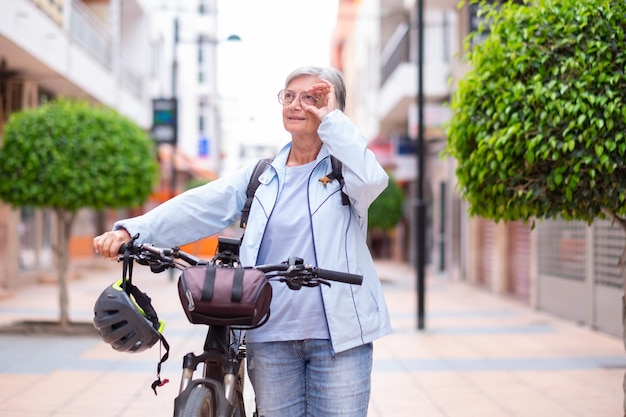 Aantrekkelijke senior wielrenner vrouw die zijn elektrische fiets duwt in een stadsstraat en omhoog kijkt Oudere actieve grootmoeder die geniet van een gezonde levensstijl en vrije tijd met pensioen