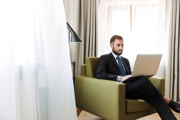 Aantrekkelijke serieuze jonge zakenman met een pak zittend in een stoel in de hotelkamer, werkend op een laptopcomputer