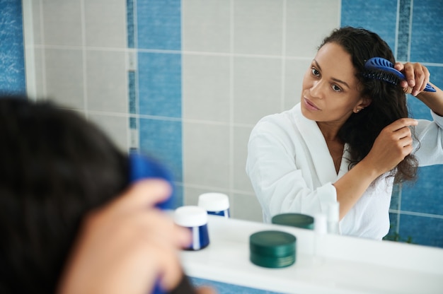 Aantrekkelijke Spaanse vrouw die een witte wafelbadjas draagt en naar haar spiegelreflectie kijkt terwijl ze haar in de badkamer kamt