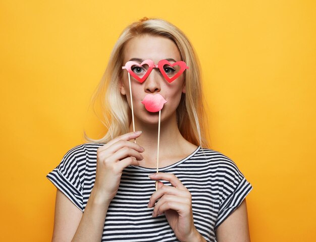 Aantrekkelijke speelse jonge vrouw met lippen en bril op een stokje over gele achtergrond