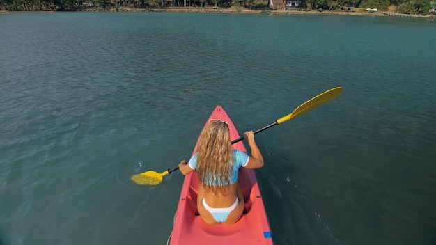 Aantrekkelijke sportieve blonde vrouw met zonnebril rijd roze plastic kano langs zeewater