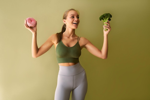 Aantrekkelijke sportieve vrouw die in de buurt van een groene achtergrond staat met een roze donut in de ene hand en broccoli in de andere, broccoli kijkend en glimlachen Keuze twijfelt aan ongezond en gezond voedsel