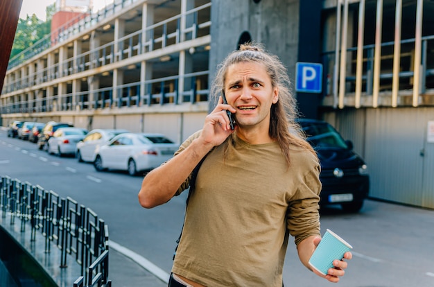 Aantrekkelijke stijlvolle hipster man drinkt koffie uit een papieren beker en praat aan de telefoon op de parkeerplaats bij de supermarkt.