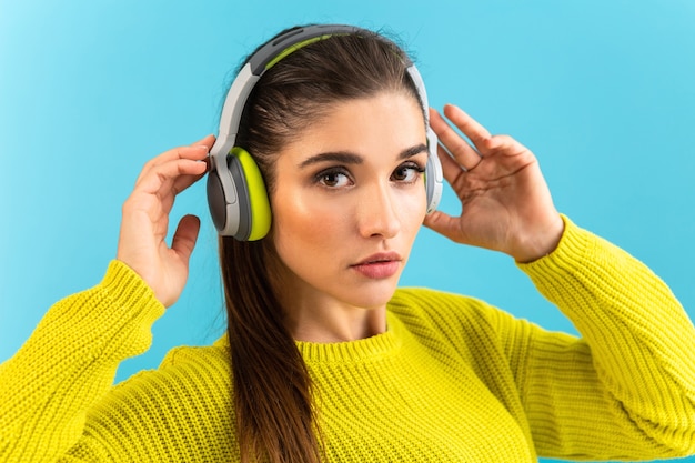 Aantrekkelijke stijlvolle jonge vrouw luisteren naar muziek in draadloze koptelefoon gelukkig dragen gele gebreide trui kleurrijke stijl mode poseren geïsoleerd op blauwe achtergrond