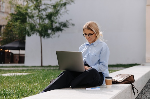 Aantrekkelijke stijlvolle vrouw in brillen typen op draagbare laptop zittend buiten. Gelukkig blondje genieten van werken op afstand aan frisse lucht.