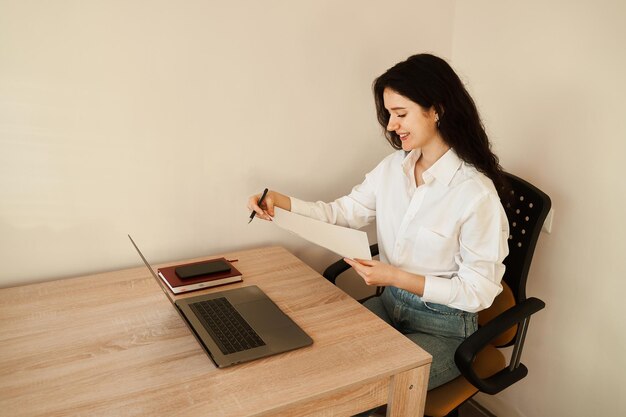 Aantrekkelijke studente die papieren vasthoudt en online studeert met behulp van laptop thuis Online onderwijs en afstandsonderwijs