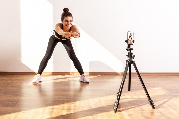 Aantrekkelijke trainer die online stretching-klasse uitzendt, video opneemt via mobiele telefoon op statief, zwarte sporttop en panty draagt. Studio-opname van volledige lengte verlicht door zonlicht vanuit het raam.