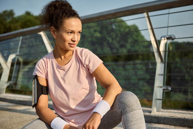 Aantrekkelijke vastberaden Afro-Amerikaanse vrouwelijke atleet, mooie sportvrouw in roze t-shirt wegkijkend zittend op een gehurkte positie op de glazen stadsbrug, genietend van zonnige ochtendtraining