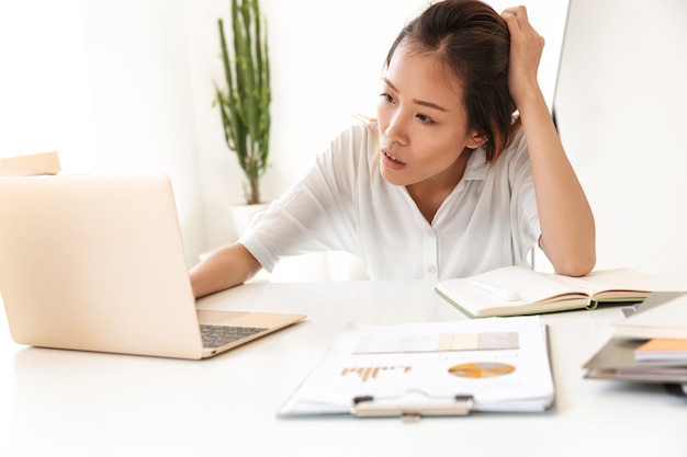 Aantrekkelijke vermoeide jonge aziatische zakenvrouw die werkt terwijl ze aan de balie op kantoor zit en op een laptop werkt