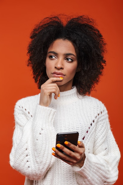 Aantrekkelijke verwarde jonge afrikaanse vrouw die een sweater draagt die mobiele telefoon houdt die over rode muur wordt geïsoleerd