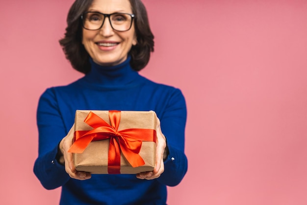 Aantrekkelijke volwassen oude vrouw met cadeaus geschenkdoos voelt gelukkige studio geïsoleerd over roze achtergrond. Levensgebeurtenissen viering felicitatie liefde zorg concept