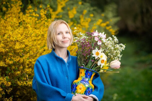 Aantrekkelijke volwassen vrouw in de herfst of lente in een blauwe jas met een boeket van verschillende bloemen tegen de achtergrond van een bloeiende gele struik