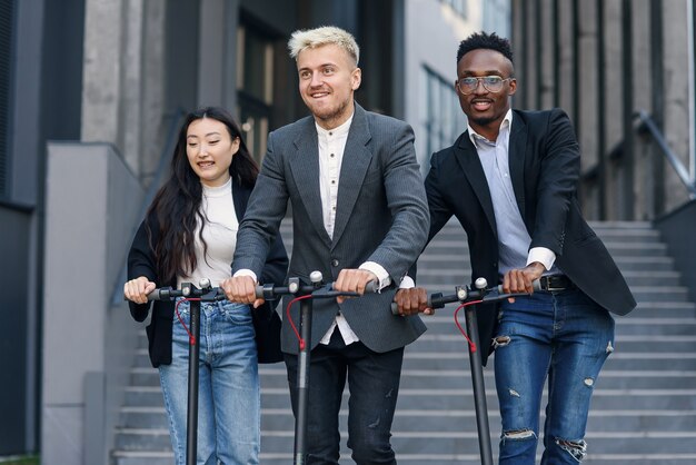 Aantrekkelijke vreugdevolle multiraciale kantoorcollega's rijden op elektrische scooters in de buurt van een prachtig architectonisch gebouw.