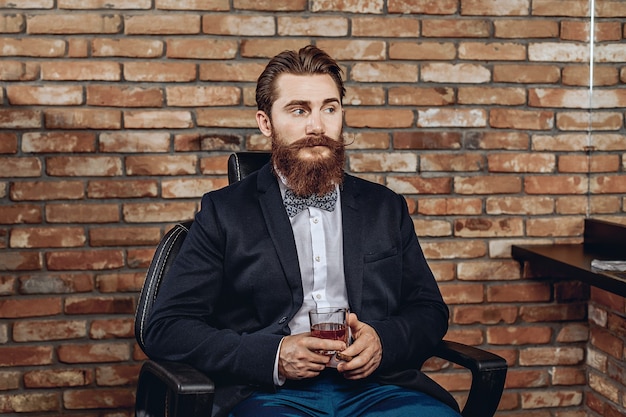 Foto aantrekkelijke vrij mooie man in een pak zittend op de stoel en met een glas whisky en een sigaar in zijn handen.