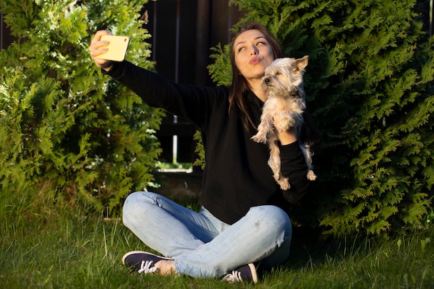 Aantrekkelijke vrolijke brunette vrouw met lang haar gekleed in zwarte hoodie maakt emotioneel kus selfies met yorkshire hond op achtergrond van groene tui